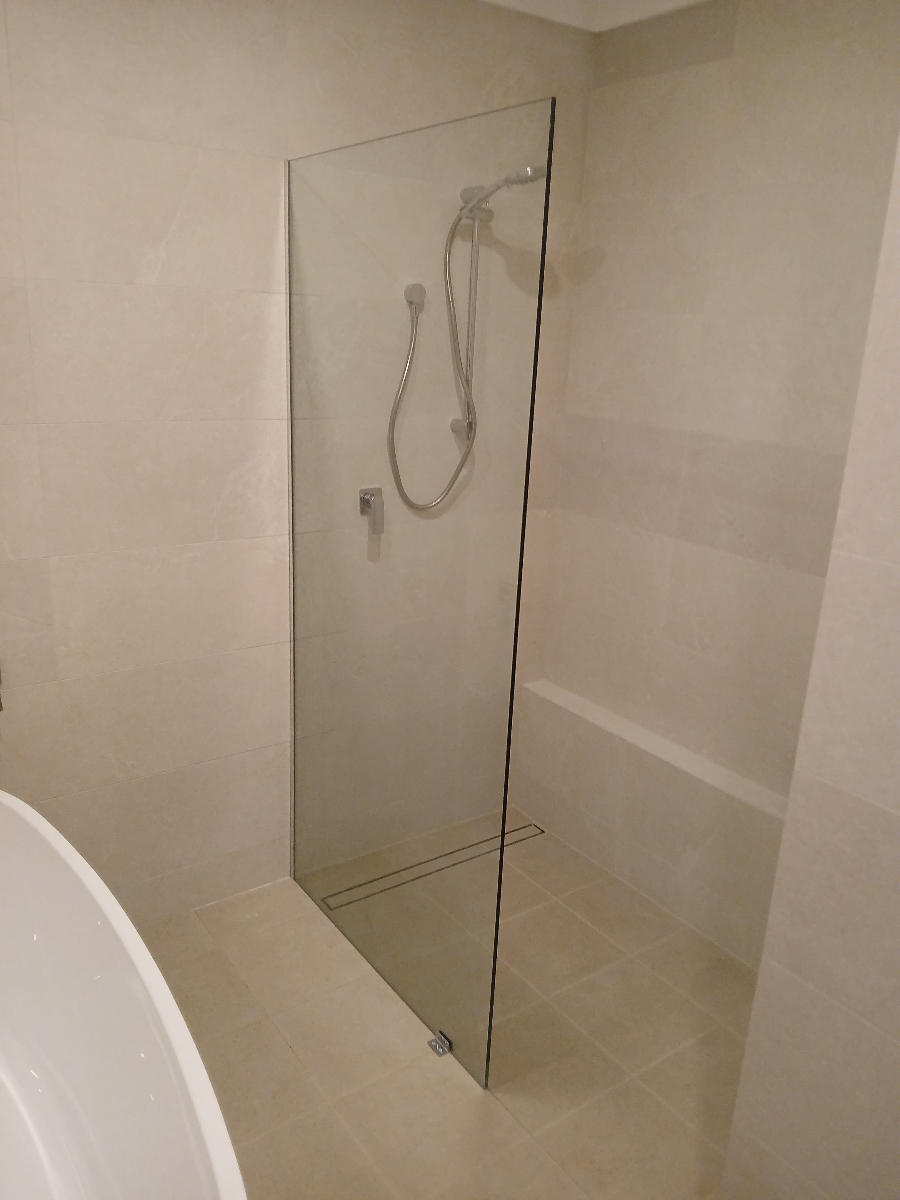 Modern walk-in shower featuring a frameless glass panel, handheld showerhead, and neutral beige tiling.