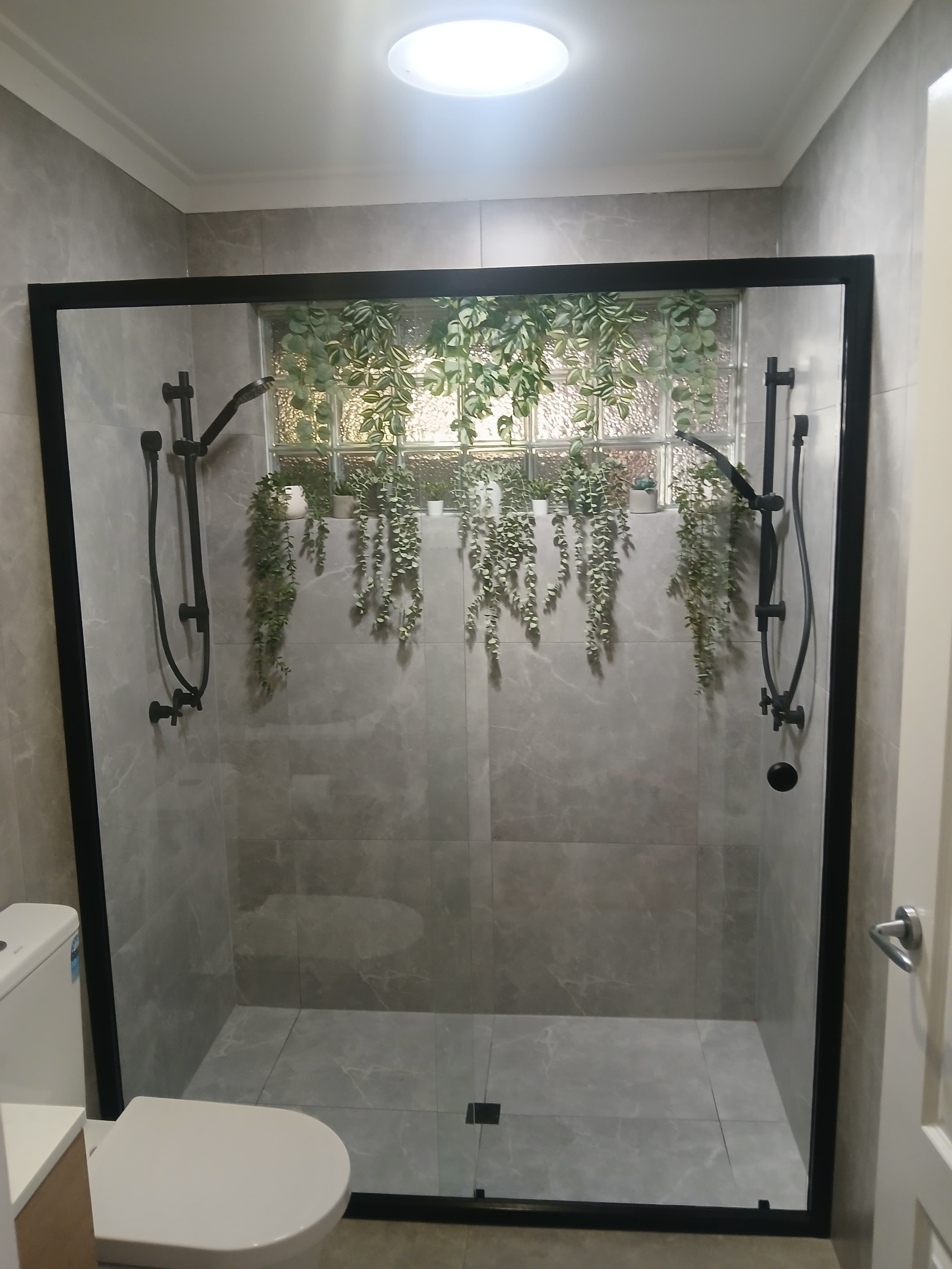 Modern shower with grey marble tiles, black fixtures, and green plants over glass blocks.