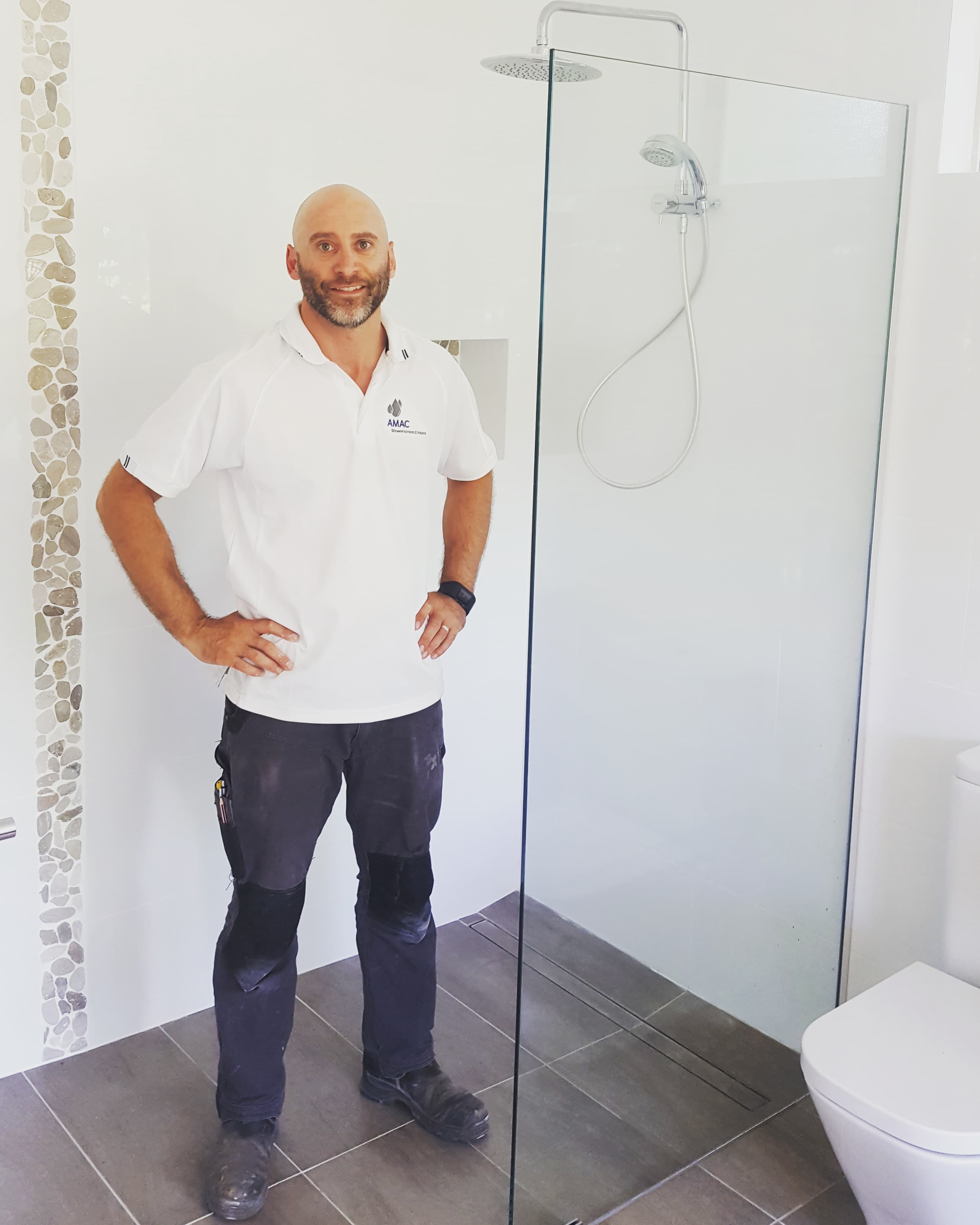 Smiling tradesman in white polo shirt standing in a modern bathroom with glass shower screen.